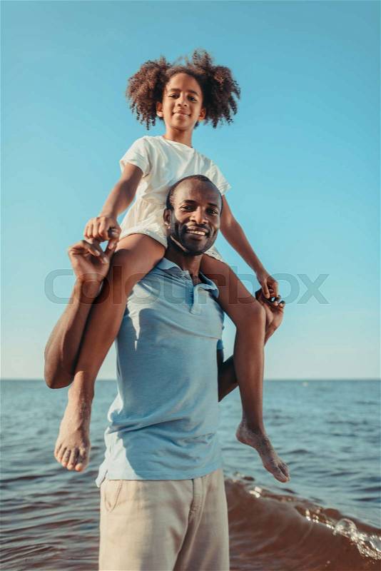 Happy african american father carrying little daughter on neck and smiling at camera on beach , stock photo