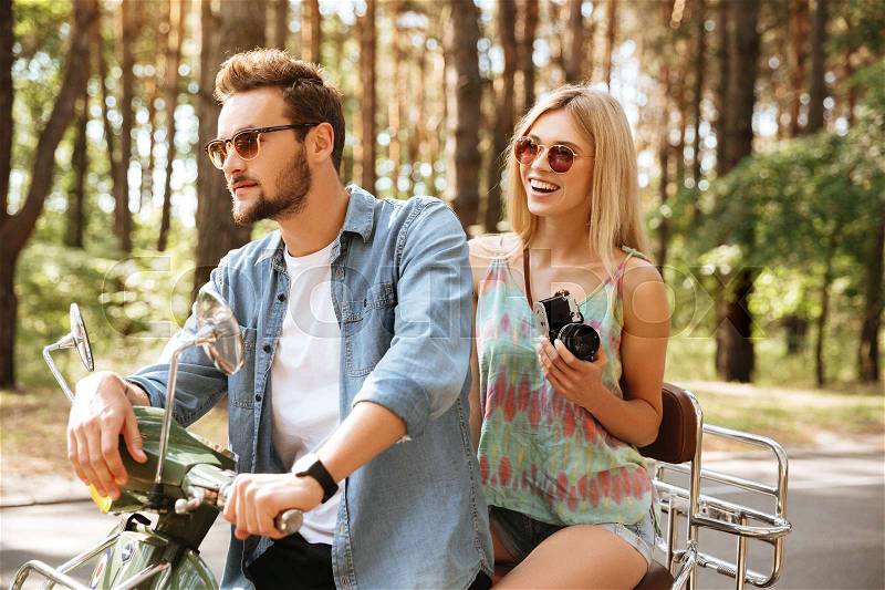 Picture of young bearded man on scooter with girlfriend holding camera outdoors. Looking aside, stock photo