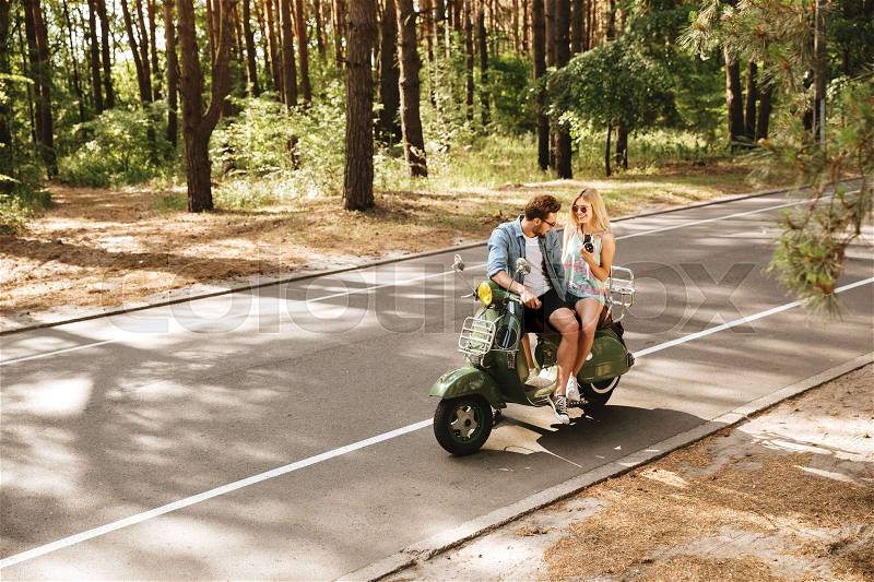 Image of young handsome man on scooter with girlfriend holding camera outdoors. Looking aside, stock photo