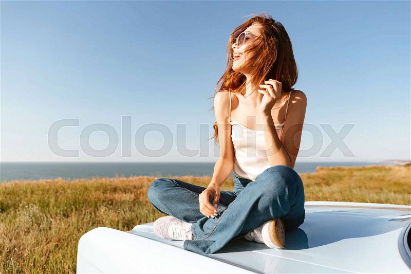 Young attractive woman relaxing while sitting on a car outdoors and looking at the landscape, stock photo