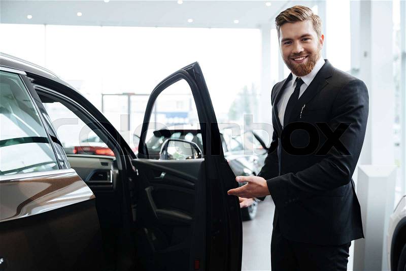 Happy smiling male dealer inviting customers to sit in a new car, stock photo