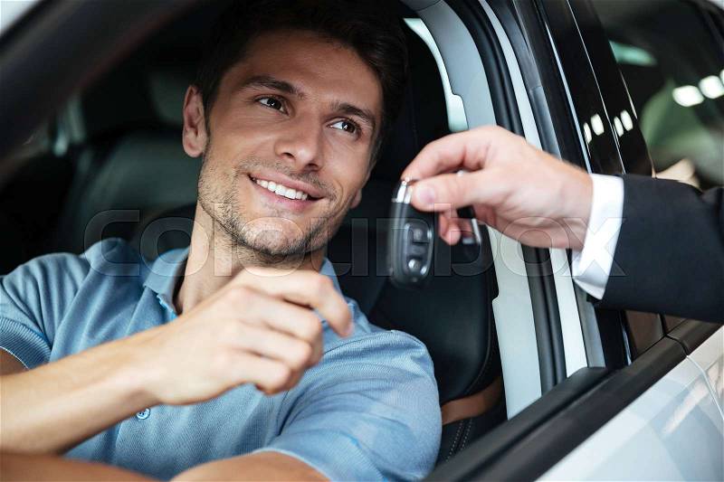 Smiling happy man sitting in his new car and taking keys from a dealers hand at the salon, stock photo
