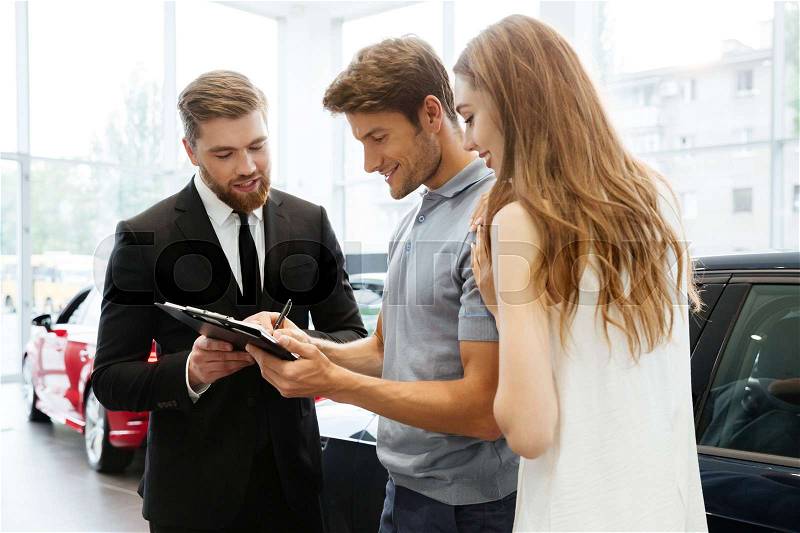 Handsome salesman filing documents with his happy clients at the dealership, stock photo