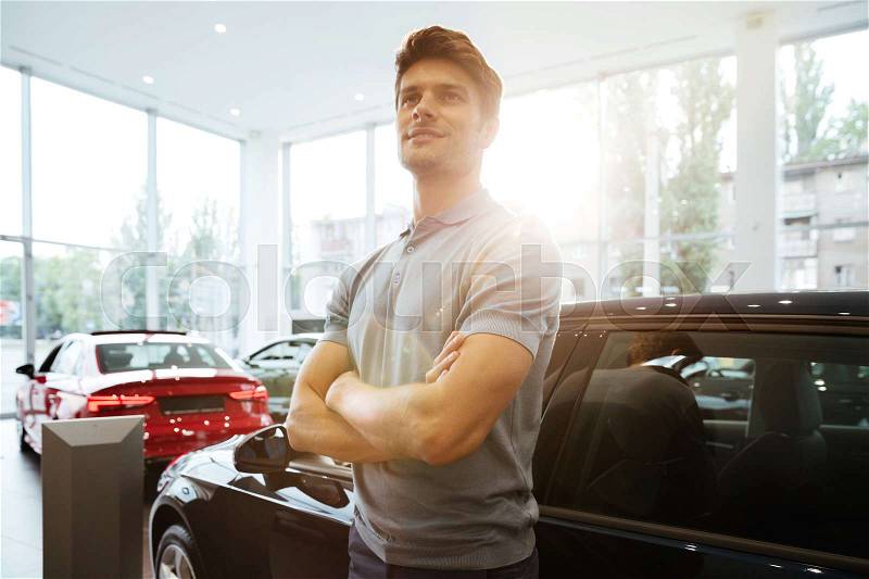 Handsome happy man standing in front of a car at the dealership center, stock photo