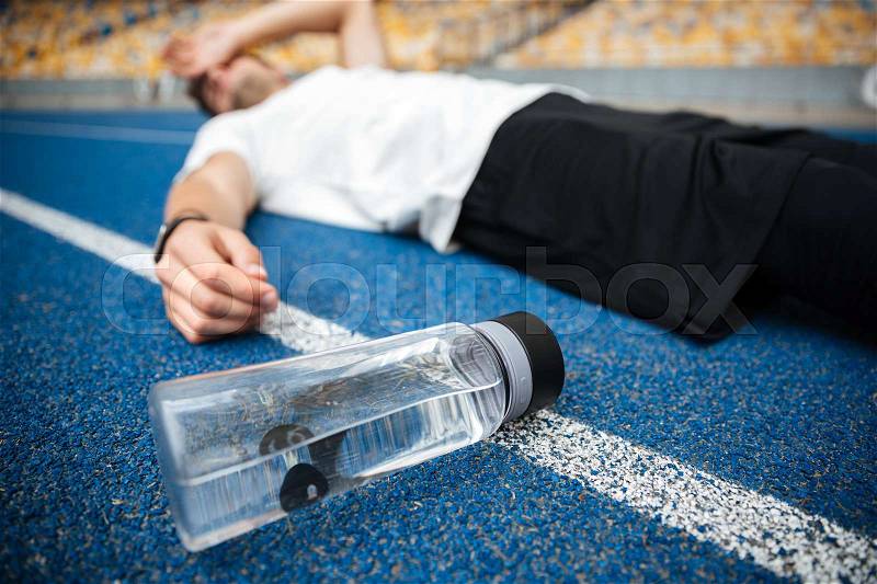 Exhausted young sportsman lying on a racetrack with bottle of water at the stadium, stock photo