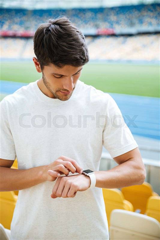 Picture of concentrated young sports man looking at watch at the stadium outdoors, stock photo