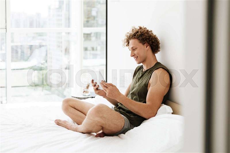 Picture of curly happy man sitting on bed at home using tablet computer. Looking aside, stock photo