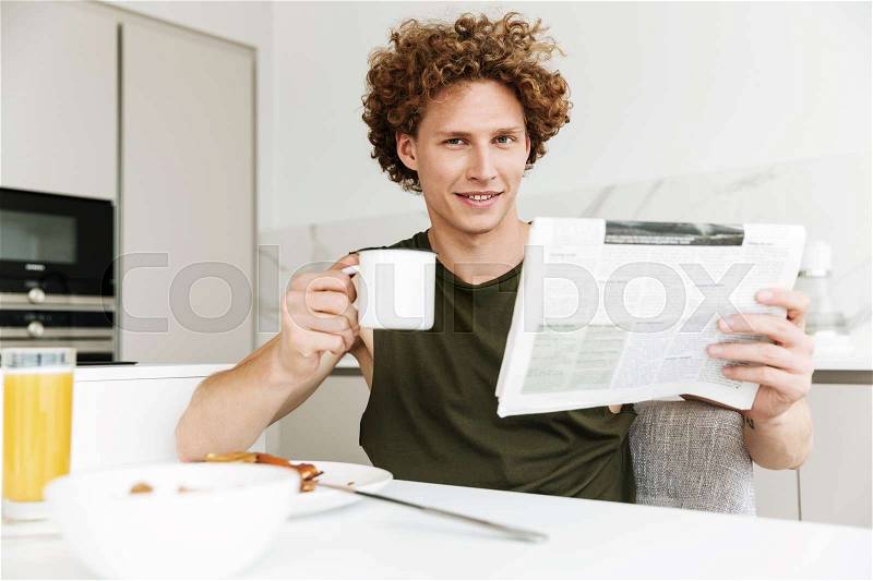 Picture of man sitting at the kitchen while holding newspaper and drinking coffee. Looking camera, stock photo