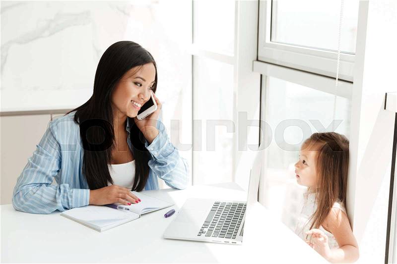 Smiling chinese woman talking on a mobile phone and working on laptop while her little daughter playing at home, stock photo