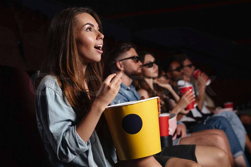 Image of young happy friends sitting in cinema watch film eating popcorn, stock photo
