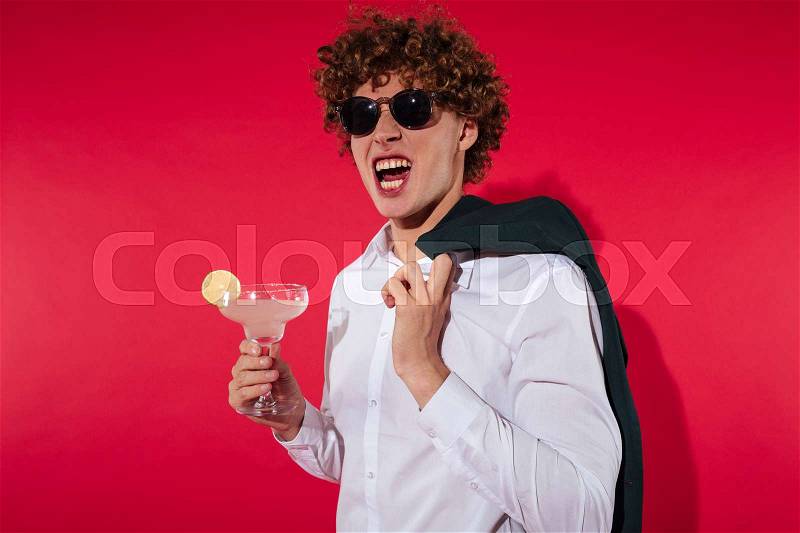 Handsome excited man in white shirt and jacket holding cocktail while standing isolated ove red background, stock photo