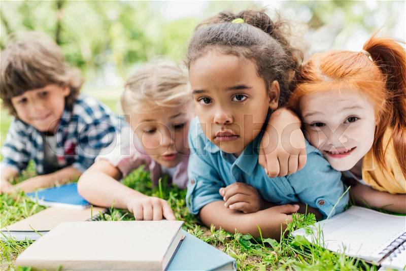 Cute multiethnic kids with books lying on grass, stock photo