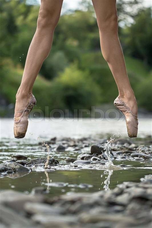 Active ballerina jumping in the shallow river on the background of the green shore. She wears beige pointes. Water splashes spreading around her legs. Closeup. Vertical, stock photo