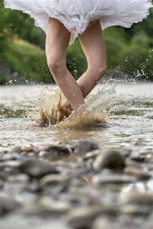 Amazing ballerina jumping in the shallow river on the background of the green shore. She wears a white tutu. Water splashes spreading around her legs. Closeup. Vertical, stock photo