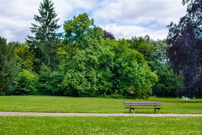 Rustic wooden slatted bench in a verdant green park with neat lawns and woodland trees at the side of a walkway or path, stock photo