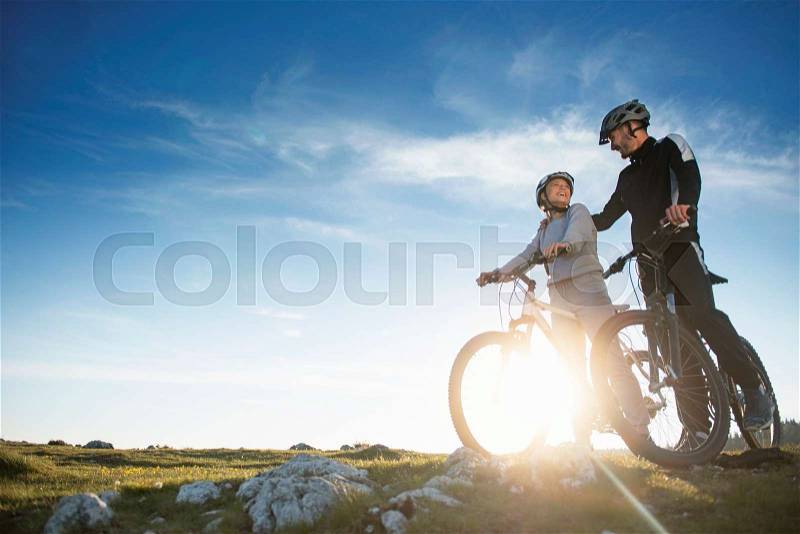 Cyclist couple with mountain bikes standing on the hill under the evening sky and enjoying bright sun at the sunset. Below is a city in the distance, stock photo
