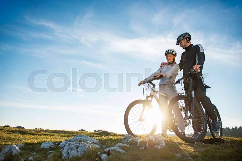 Cyclist couple with mountain bikes standing on the hill under the evening sky and enjoying bright sun at the sunset. Below is a city in the distance, stock photo