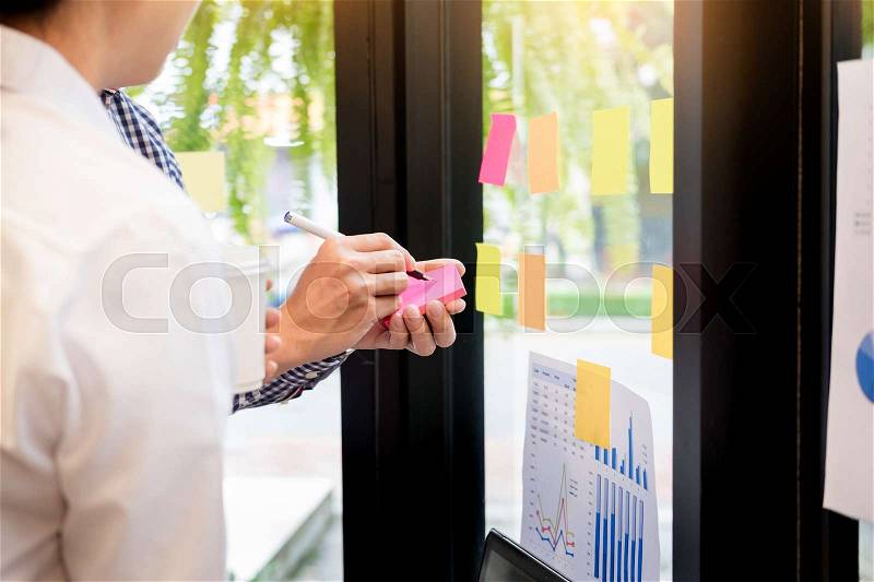 Business man sticking adhesive notes on glass wall in office and discussting with team, stock photo