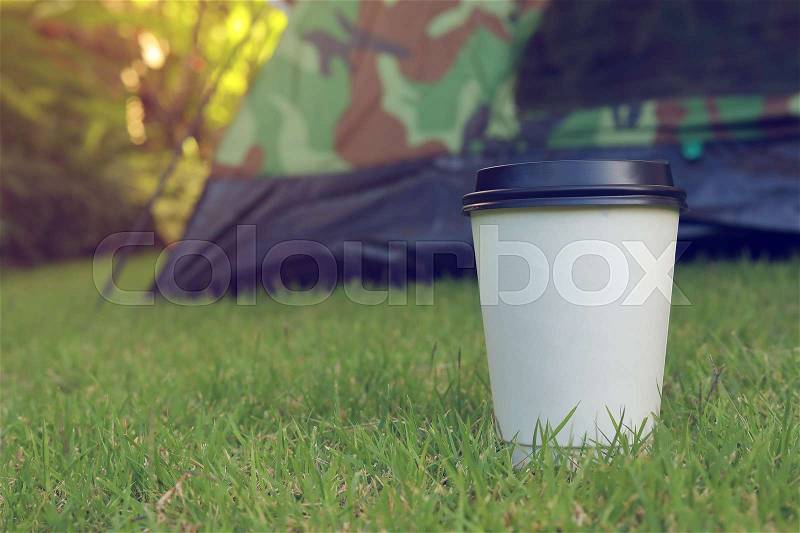 Hot coffee drink, white disposable cup on green grass lawn campsite, tent camping trip journey travel in nature, stock photo