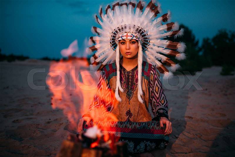 American Indian girl against bonfire in the night, female shaman, Cherokee, Navajo. Headdress made of feathers of wild birds. Traditional ritual, stock photo