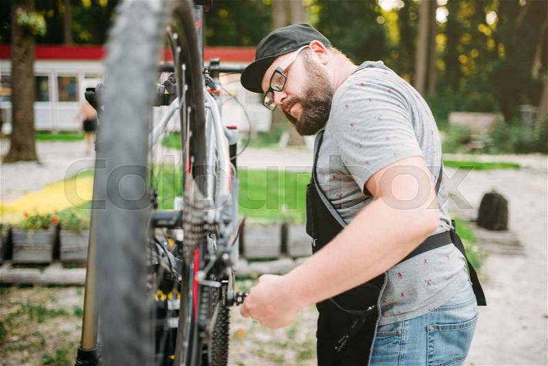 Repairman works with bike wheel, cycle workshop outdoor. Bearded bicycle mechanic in apron, stock photo