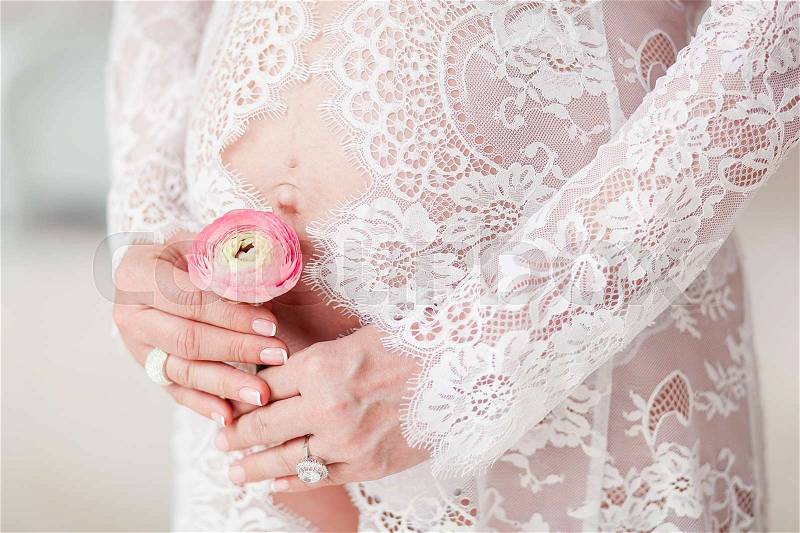 Pregnant woman holding ranunculus flower above belly in delicate lingerie, stock photo