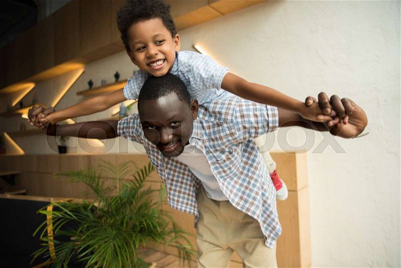 Happy african american father piggybacking adorable little son indoors, stock photo