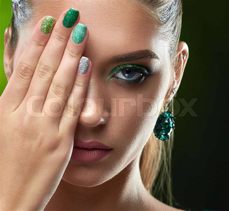 Crop of attractive brunette woman with shiny manicure, makeup in green colors, plump lips. Beautiful girl with big earring looking at camera, hiding face, showing nails. Beauty concept, stock photo