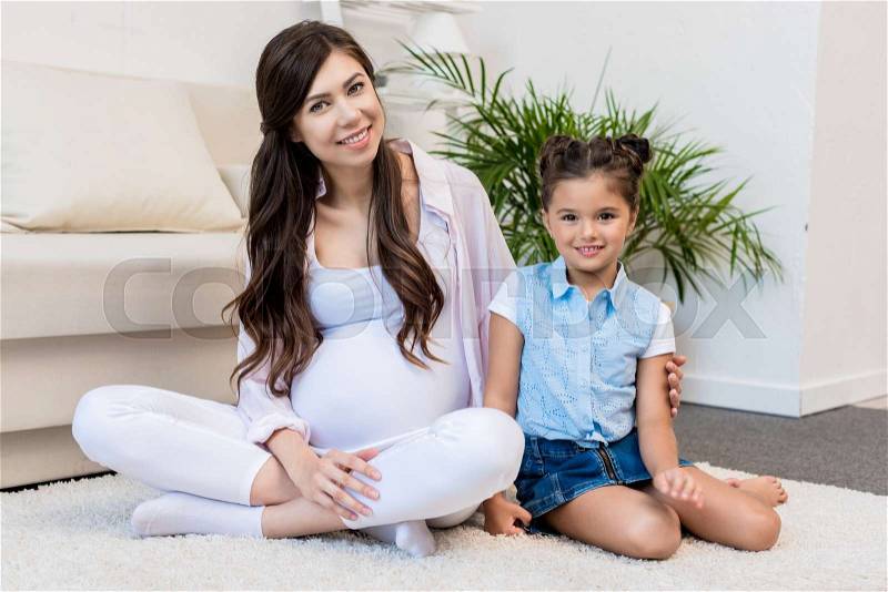 Pregnant woman sitting on a furry rug in living room with her little daughter, stock photo