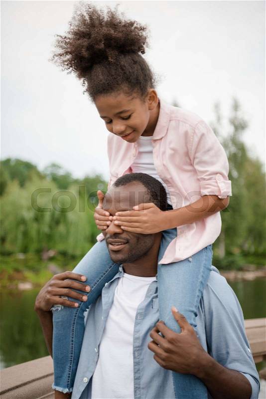 African american little girl covering fathers eyes while piggybacking together in park, stock photo