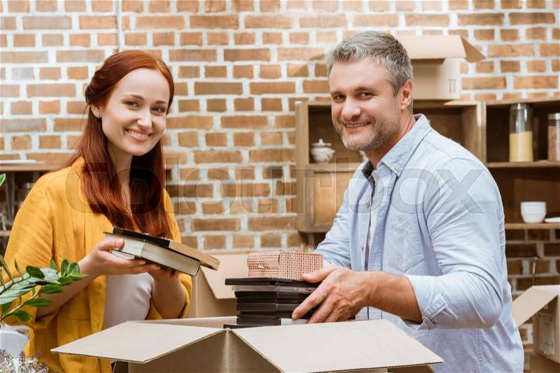 Portrait of happy couple looking at camera while unpacking box at new home, stock photo