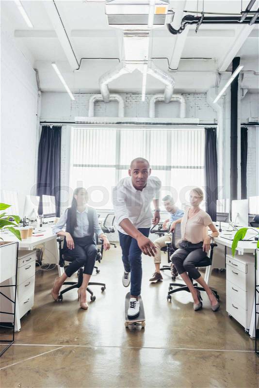 Young business people looking at african american colleague on skateboard in office, stock photo