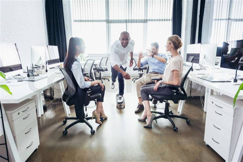 Young business people looking at african american colleague riding skateboard in office, stock photo