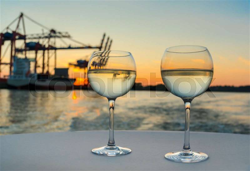 Two glasses of cool white wine on white tablecloth with harbor structures and cargo ship in blurred background at sunset, stock photo