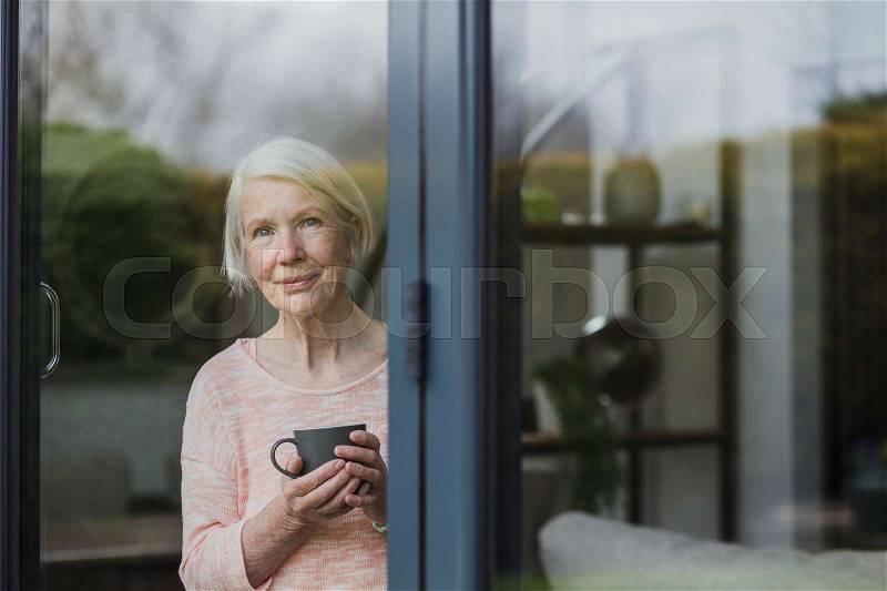 Senior woman is standing at the window in her home, enjoying a cup of tea, stock photo