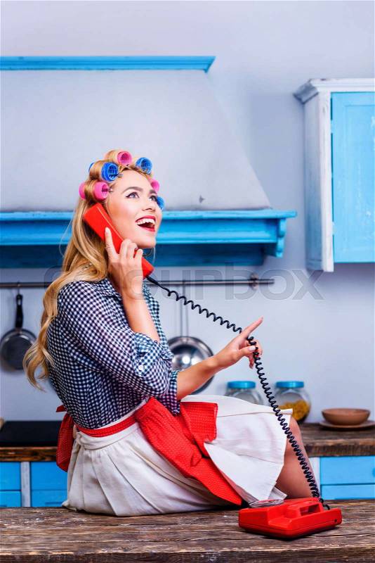 Beautiful happy woman in curlers talking on vintage telephone in kitchen , stock photo