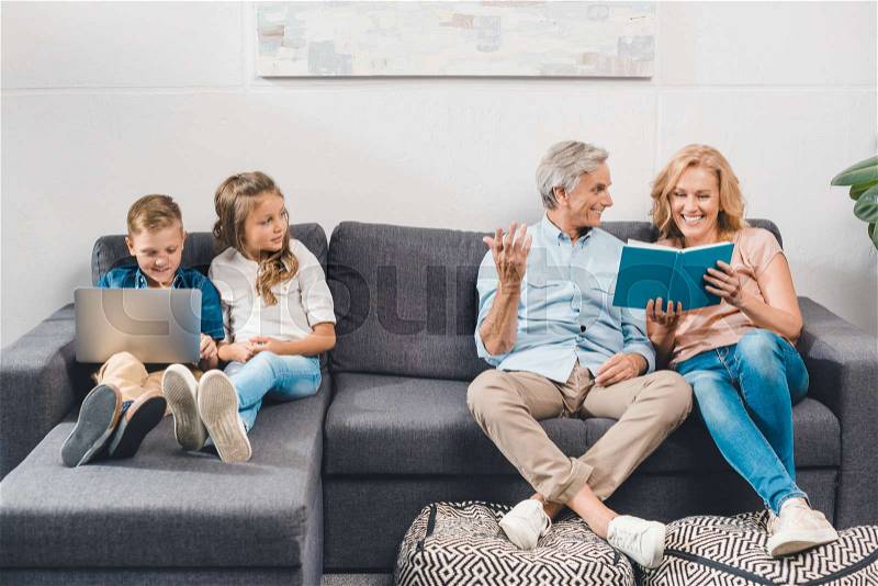 Grandparents reading book while grandchildren using laptop and sitting on sofa, stock photo