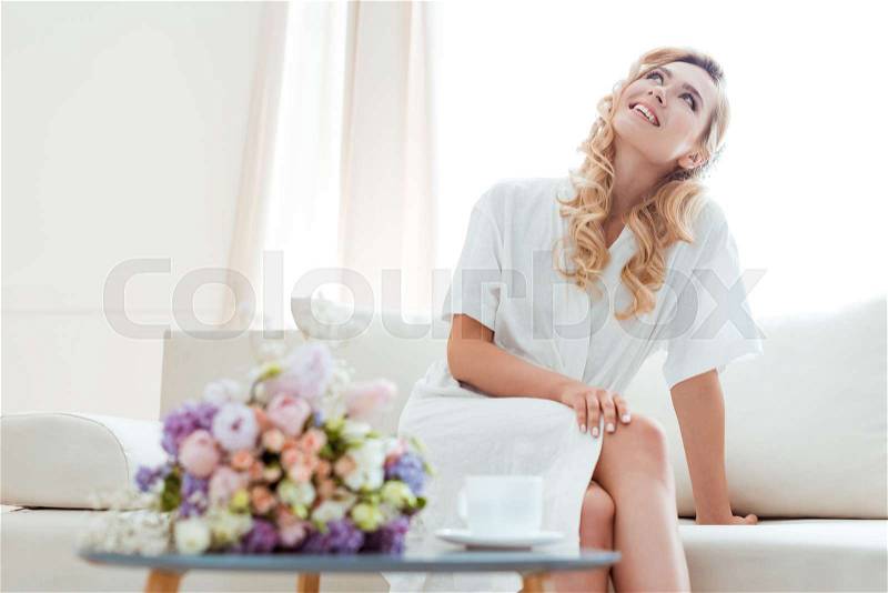 Dreamy young woman sitting on sofa with cup of coffee and wedding bouquet and looking up, stock photo