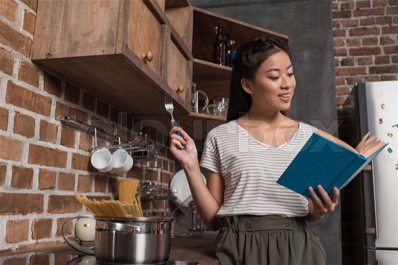 Asian female student reading book while cooking in kitchen, stock photo