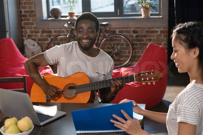 African american male student playing guitar while happy asian girl studying with copybook near, stock photo