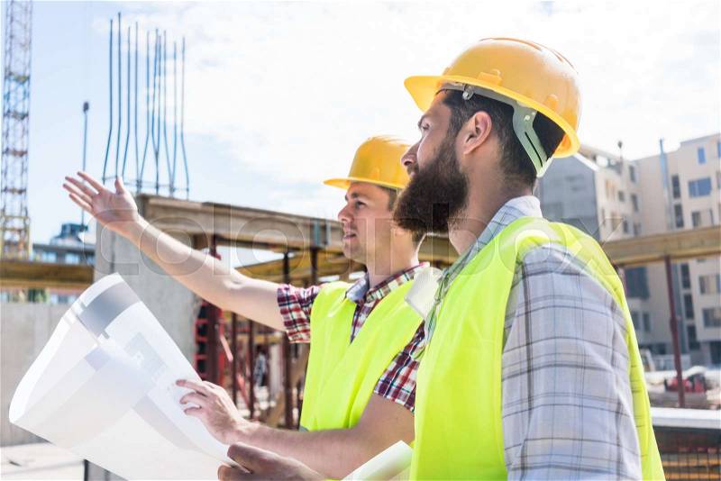 Side view of a skilled worker sharing with his colleague his innovative vision about a building while holding a blueprint outdoors on the construction site , stock photo