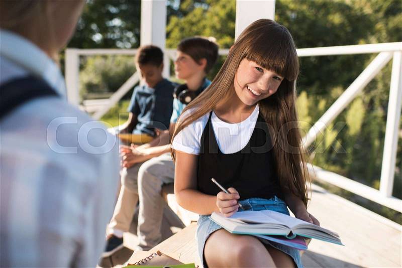 Beautiful smiling teenage girl studying while classmates sitting behind outdoors, stock photo