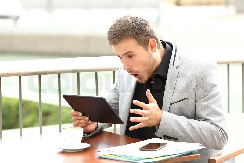 Single amazed executive male reading surprising news in a tablet sitting in a coffee shop, stock photo
