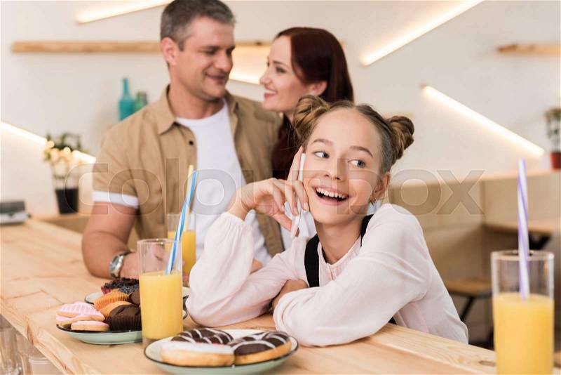 Teen girl talking by phone in cafe with her parents at bar counter, stock photo