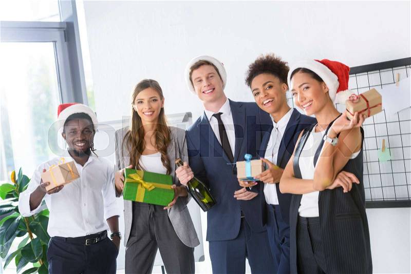 Happy young multiethnic business colleagues holding gifts and smiling at camera in office , stock photo