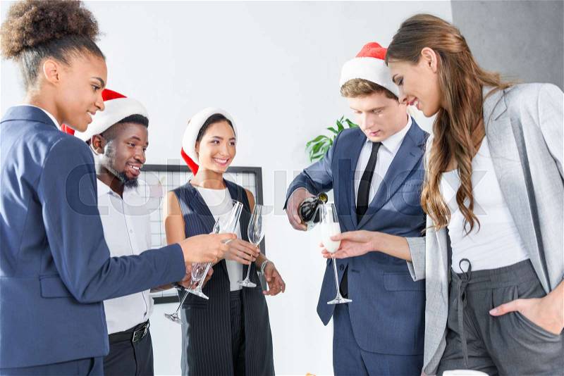 Happy young multiethnic business colleagues in santa hats drinking champagne in office , stock photo