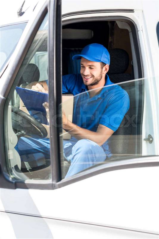 Smiling delivery man filling in documents on clipboard at car, stock photo