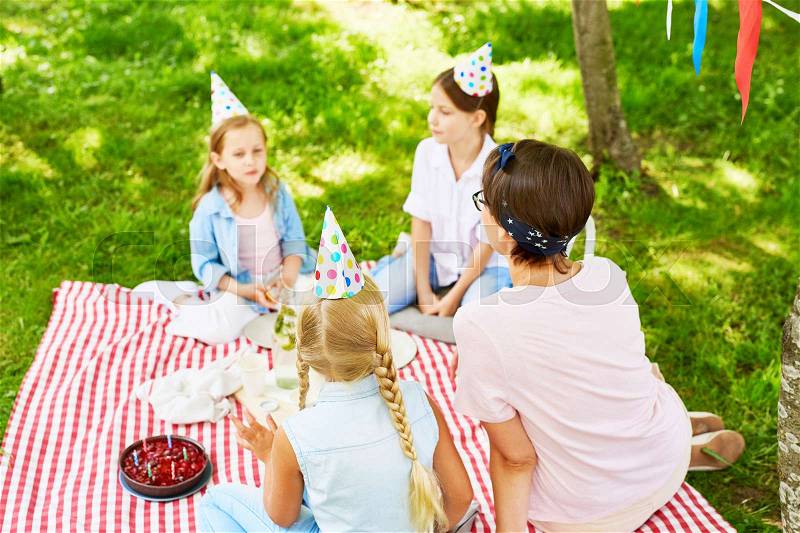 Teaher and three girls in birthday caps relaxing on green grass on summer day, stock photo
