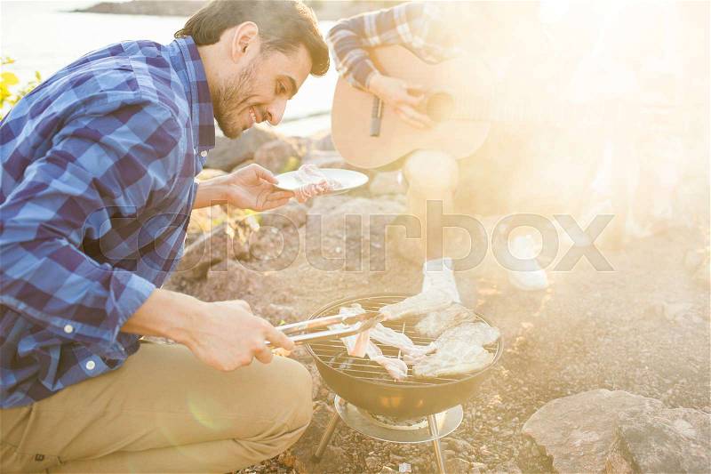 Young camper cooking grilled meat at picnic on summer weekend, stock photo