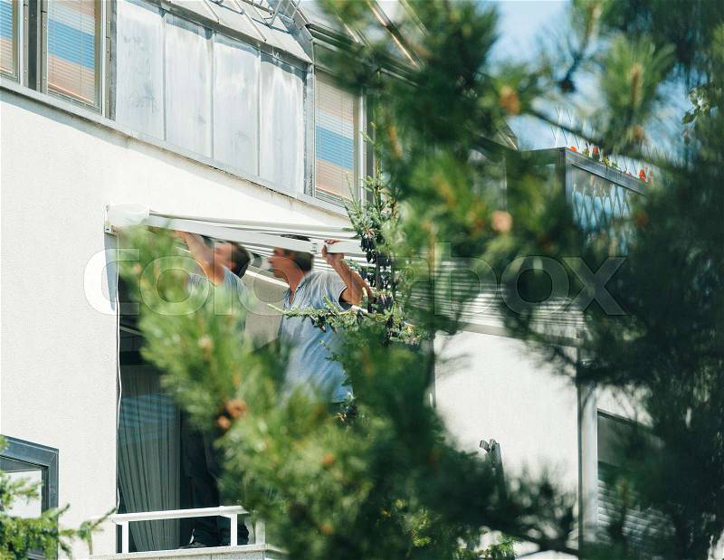 Male team installing sun protection awning for sunshade on apartment window - real life scene view through fir tree, stock photo